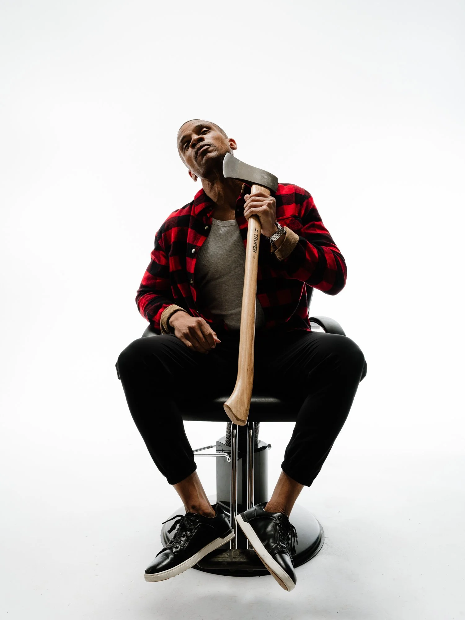 Man sitting on an office chair, holding an axe, wearing a red and black checkered shirt, black pants, and sneakers, with a blank white background.