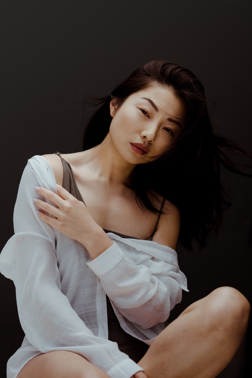 A woman with long dark hair, wearing a black tank top, white shirt, and gray bra, sitting in front of a dark background, looking at the camera with a relaxed expression.