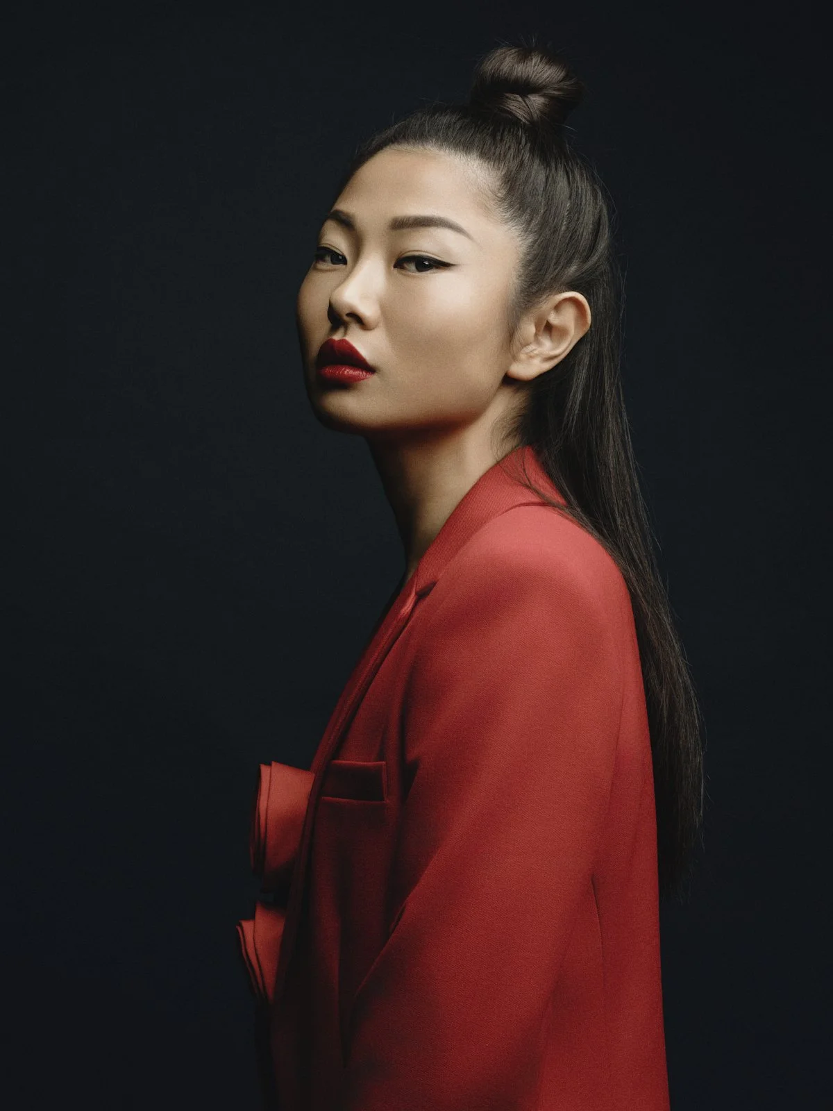 A woman with dark hair in a top bun wearing red lipstick and a red blazer against a black background.