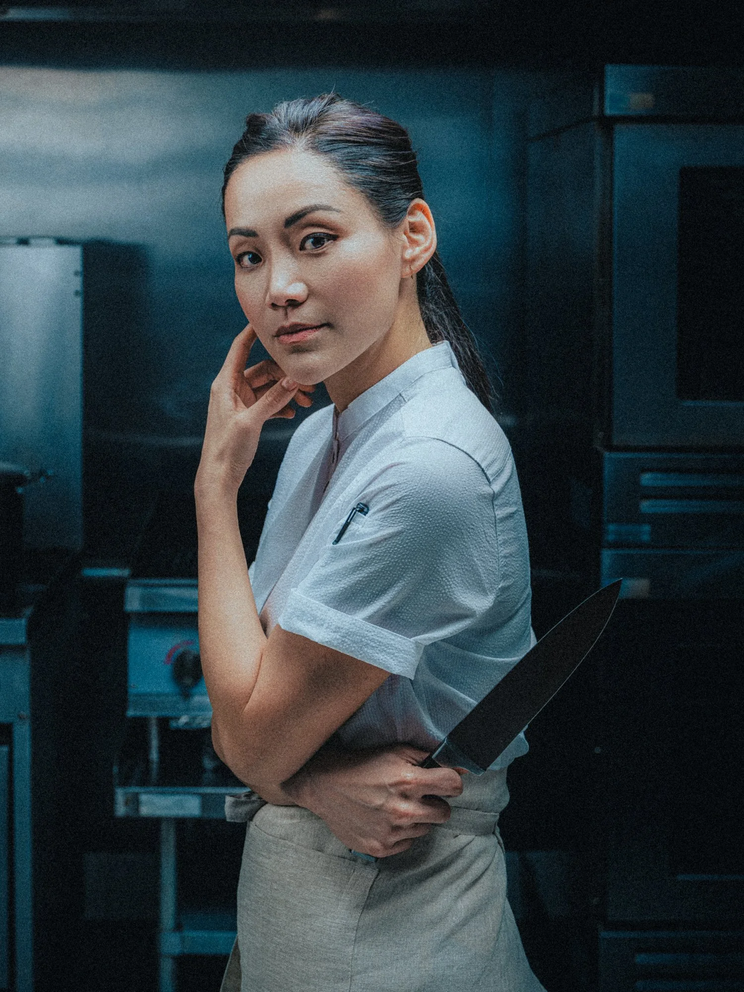 A woman with dark hair in a ponytail wearing a white blouse and beige pants, standing in a kitchen with her hand on her hip and looking at the camera.