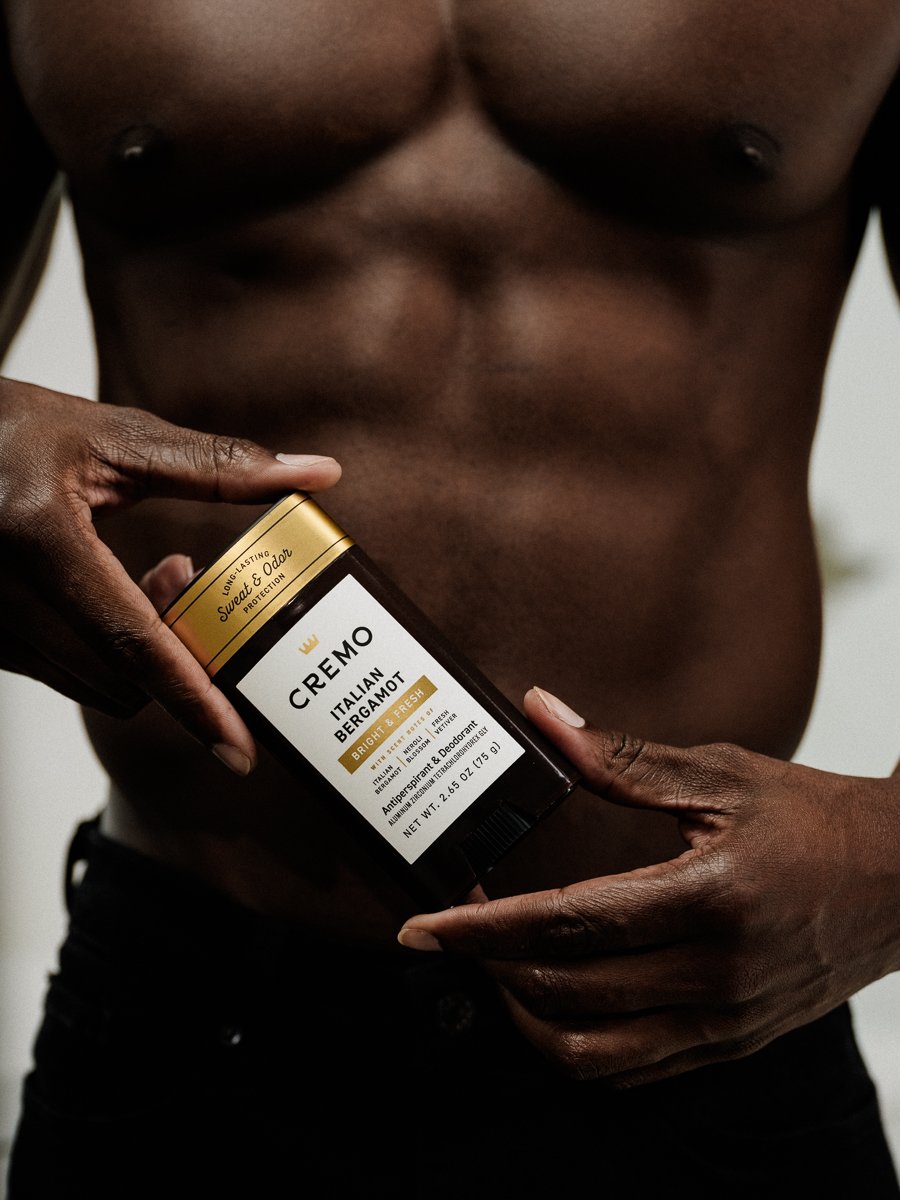 A shirtless person with dark skin holding a container of Cremo Italian Bergamot deodorant, with a plain background.