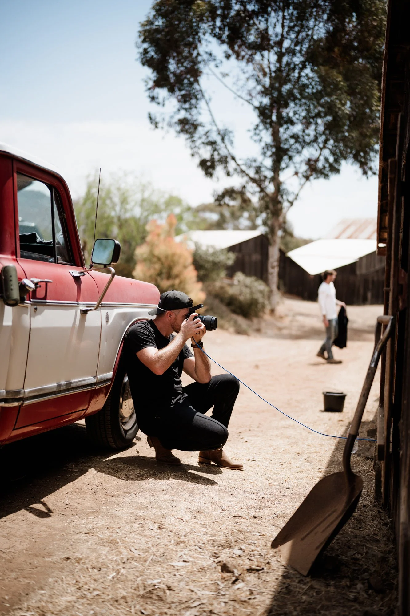 A man kneeling and taking a photograph next to a vintage red and white pickup truck, with a woman walking in the background in a rural setting with trees and barn structures.