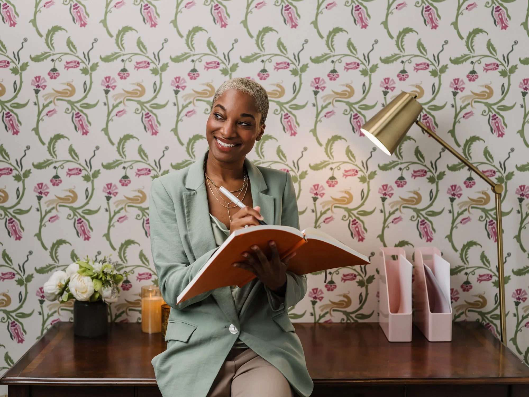 A woman with short blonde hair smiling while holding an open book or planner, sitting on a wooden table in a room with floral wallpaper background. There is a black vase with white flowers, a lit candle, and a set of pink and white magazine holders o