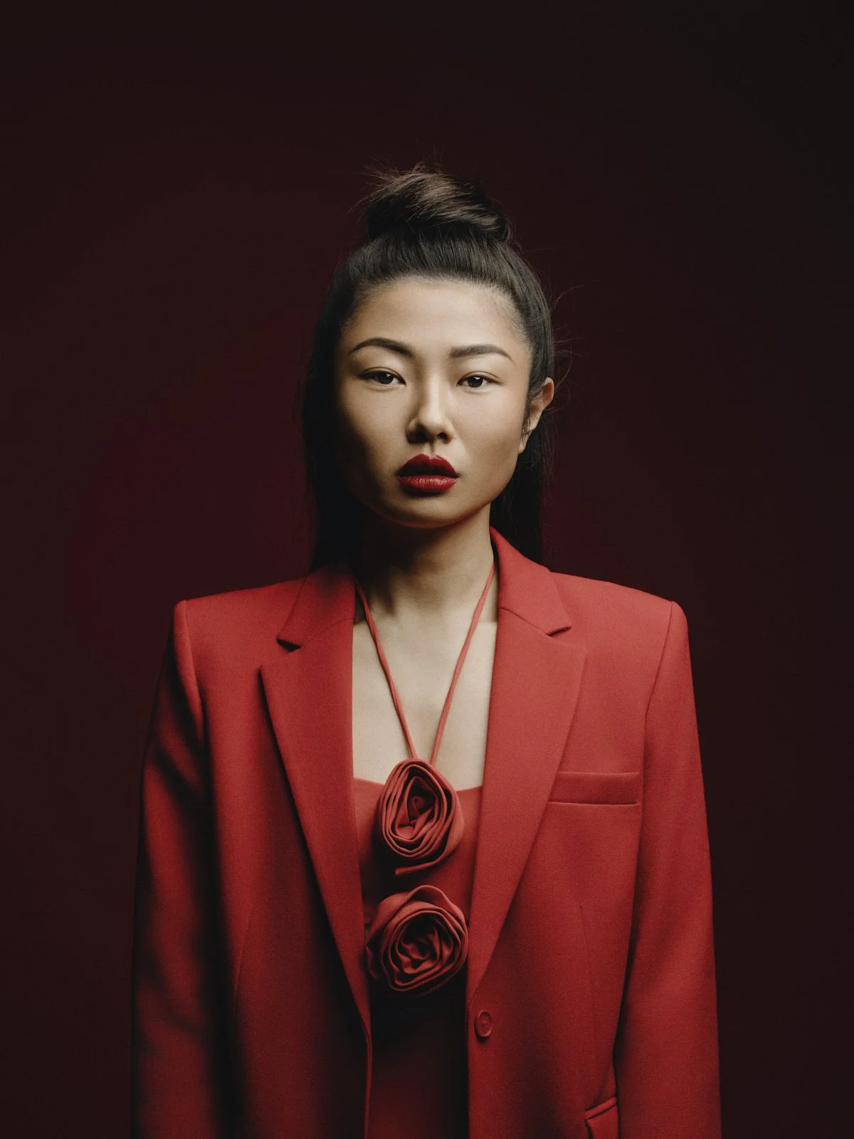 Portrait of a woman with Asian features wearing a red blazer and matching top, with a red flower necklace, against a dark red background.