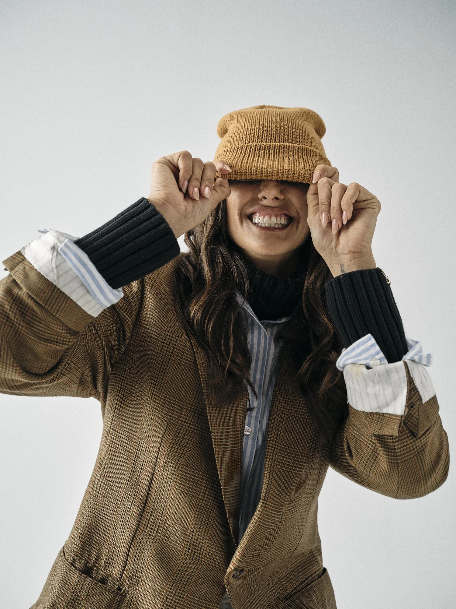 Woman wearing a tan beanie hat, smiling, holding the hat with both hands, dressed in a brown plaid blazer with a black and white ribbed sweater underneath, against a plain white background.