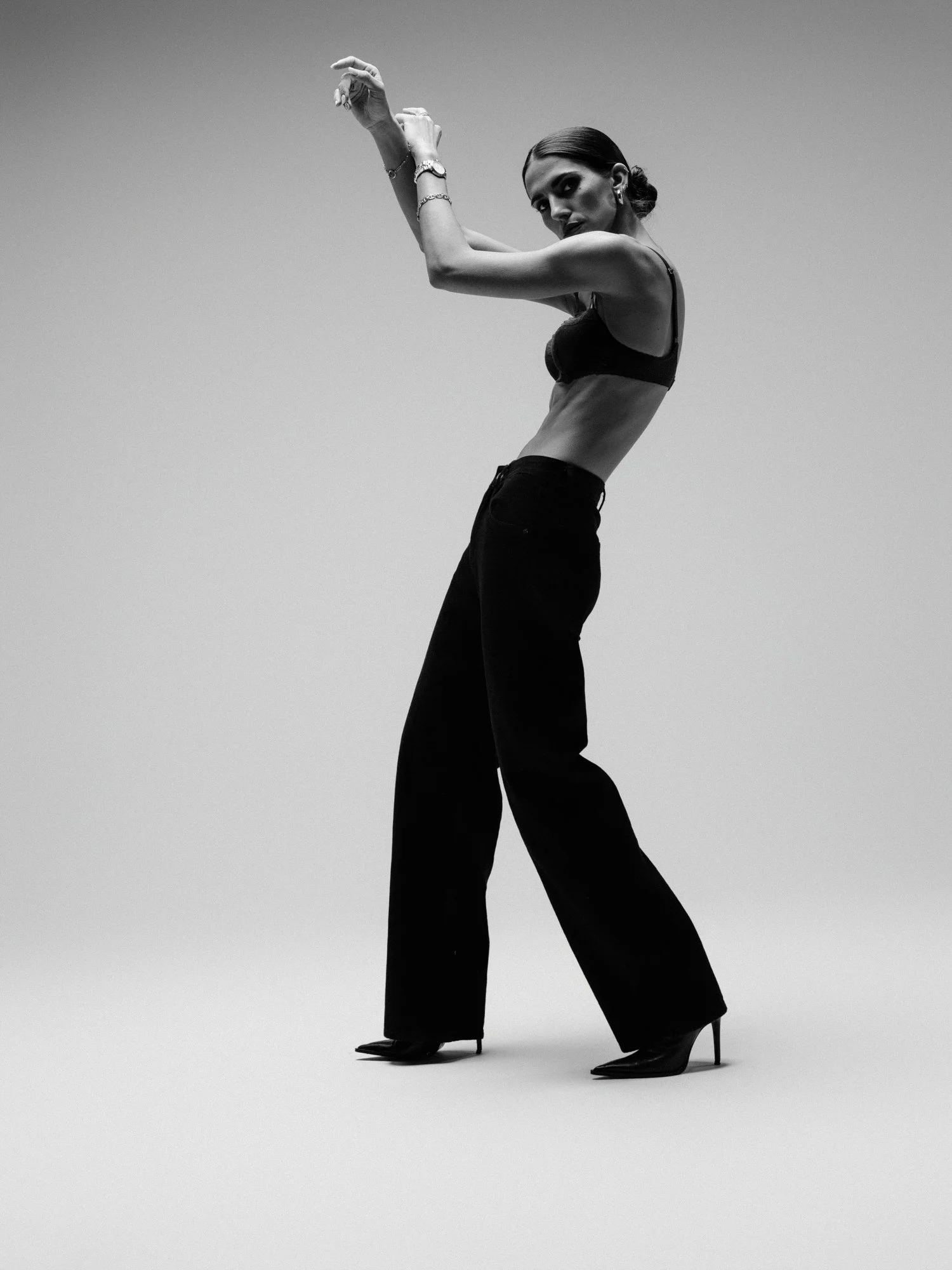Black and white photo of a woman in high heels, black bra, and black pants posing with arms raised and bent, with a clear sky background.