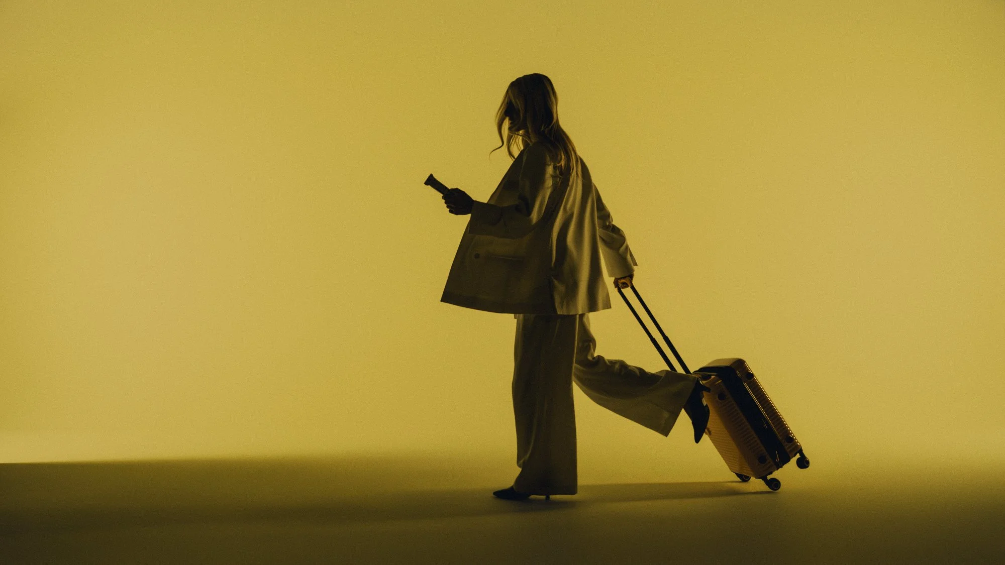 Silhouette of a woman walking with a suitcase, holding a phone, wearing a trench coat, against a yellow background, with additional luggage attached to her foot.