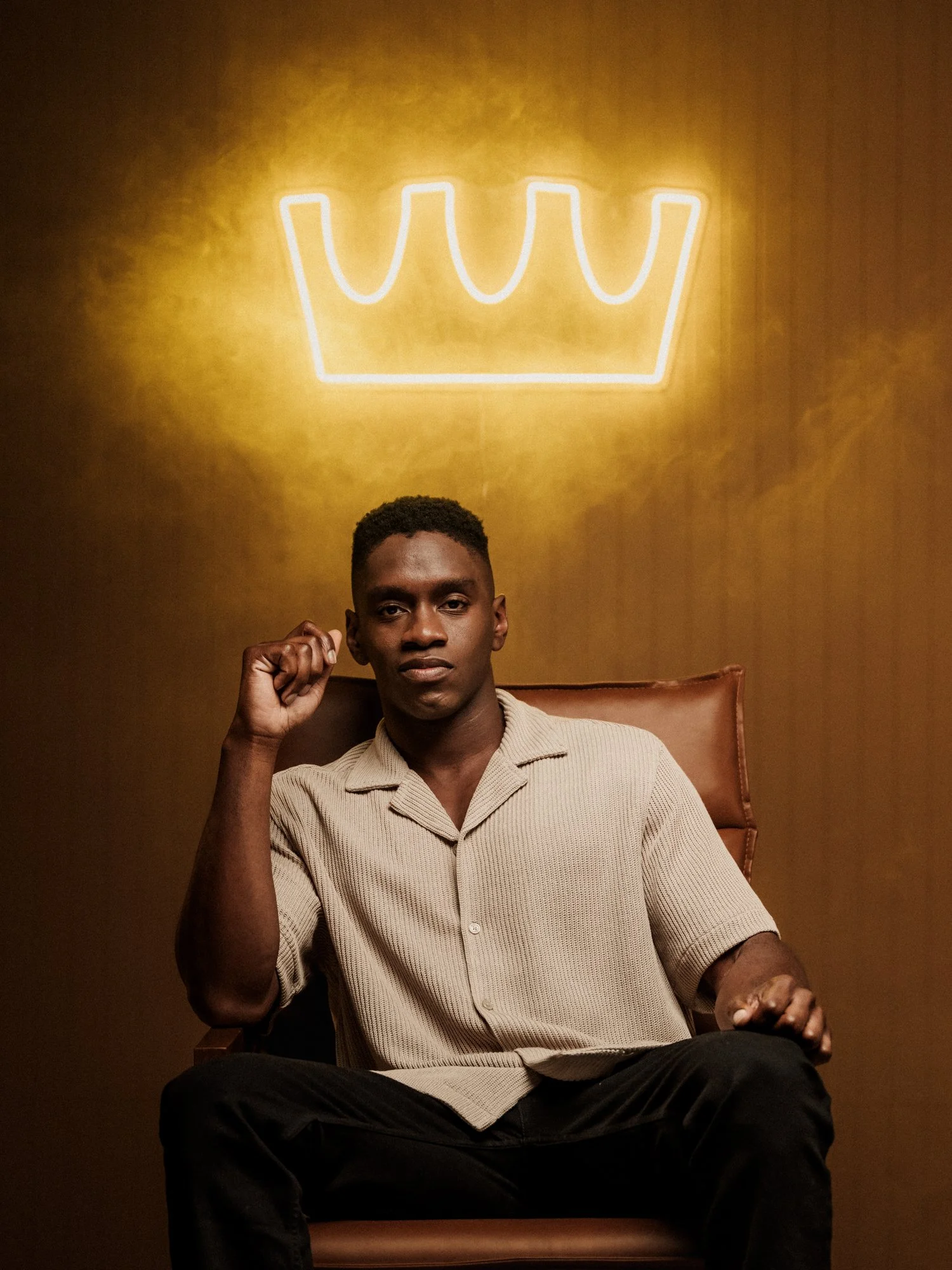 A young man sitting in a brown chair against a dark yellow wall with a neon crown sign above his head, wearing a beige short-sleeved shirt and black pants, with raised fists.