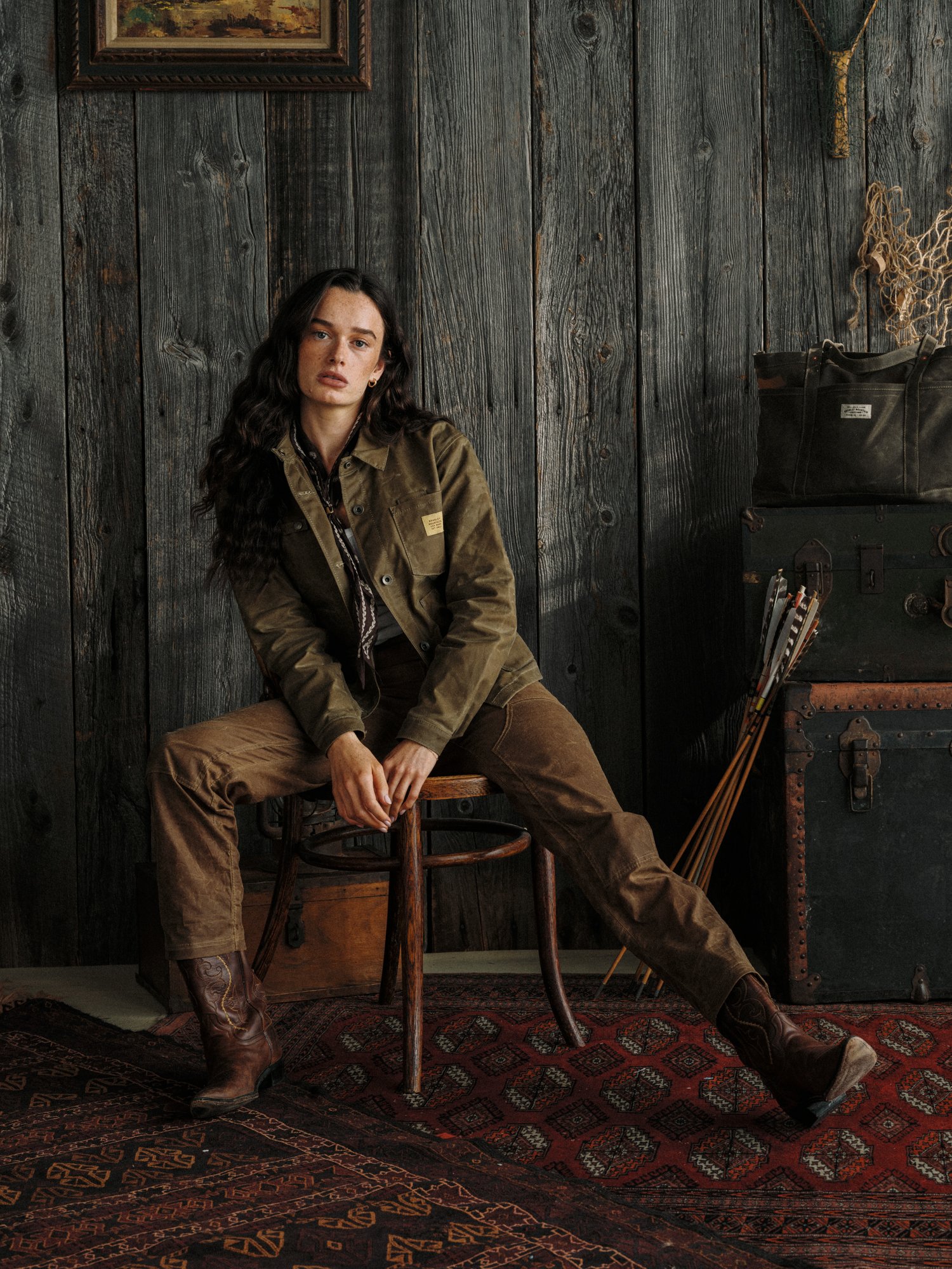 A woman with long dark hair sitting on a wooden chair against a dark wood-paneled wall. She is wearing a brown jacket, brown pants, and cowboy boots. There are vintage trunks, a bag, and a riding crop beside her. A painted landscape is framed on the 