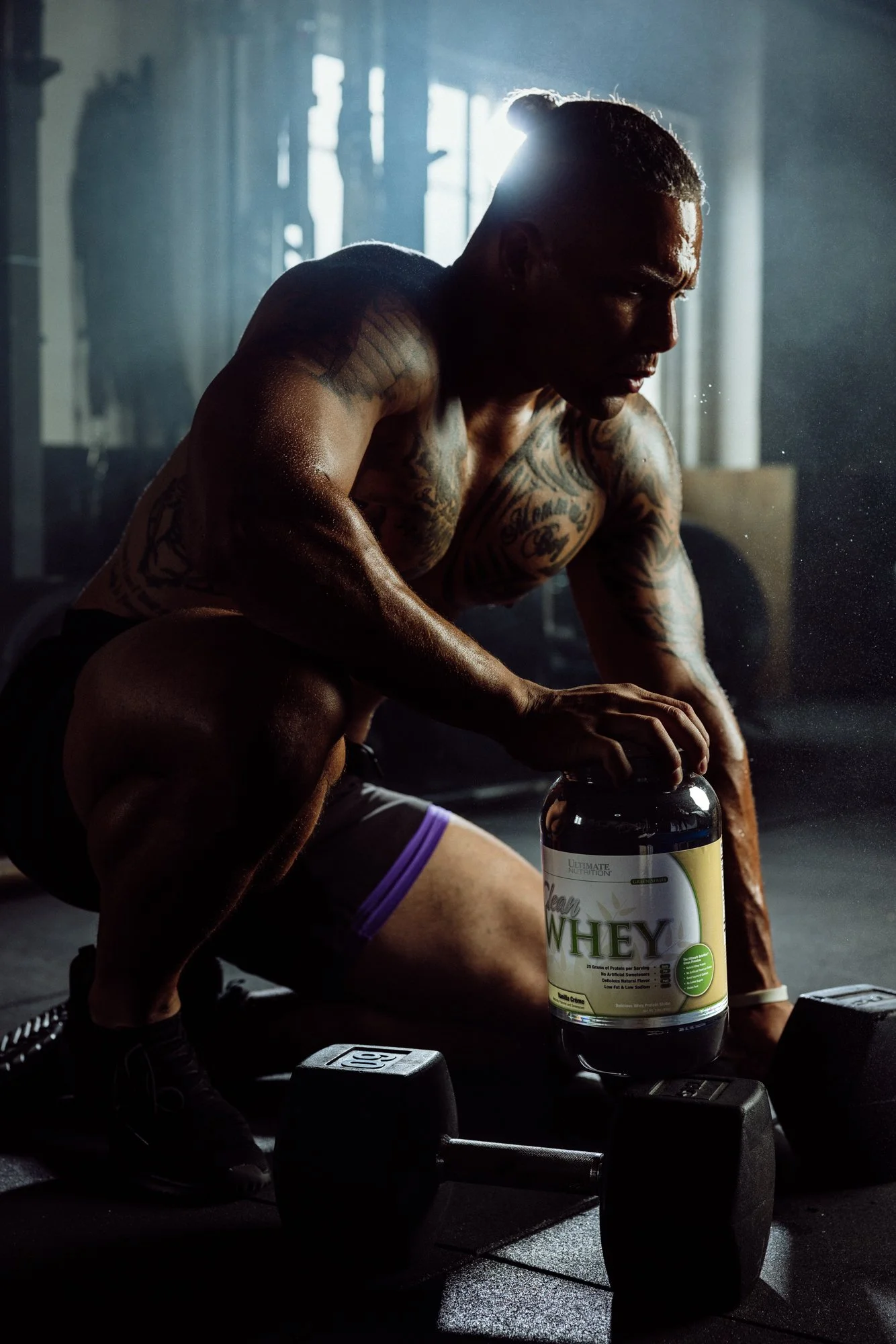 A man with tattoos wearing workout shorts and sneakers is in a gym, lifting a large container of whey protein powder, with dumbbells on the floor, and the gym appears dimly lit with sunlight streaming through windows in the background.