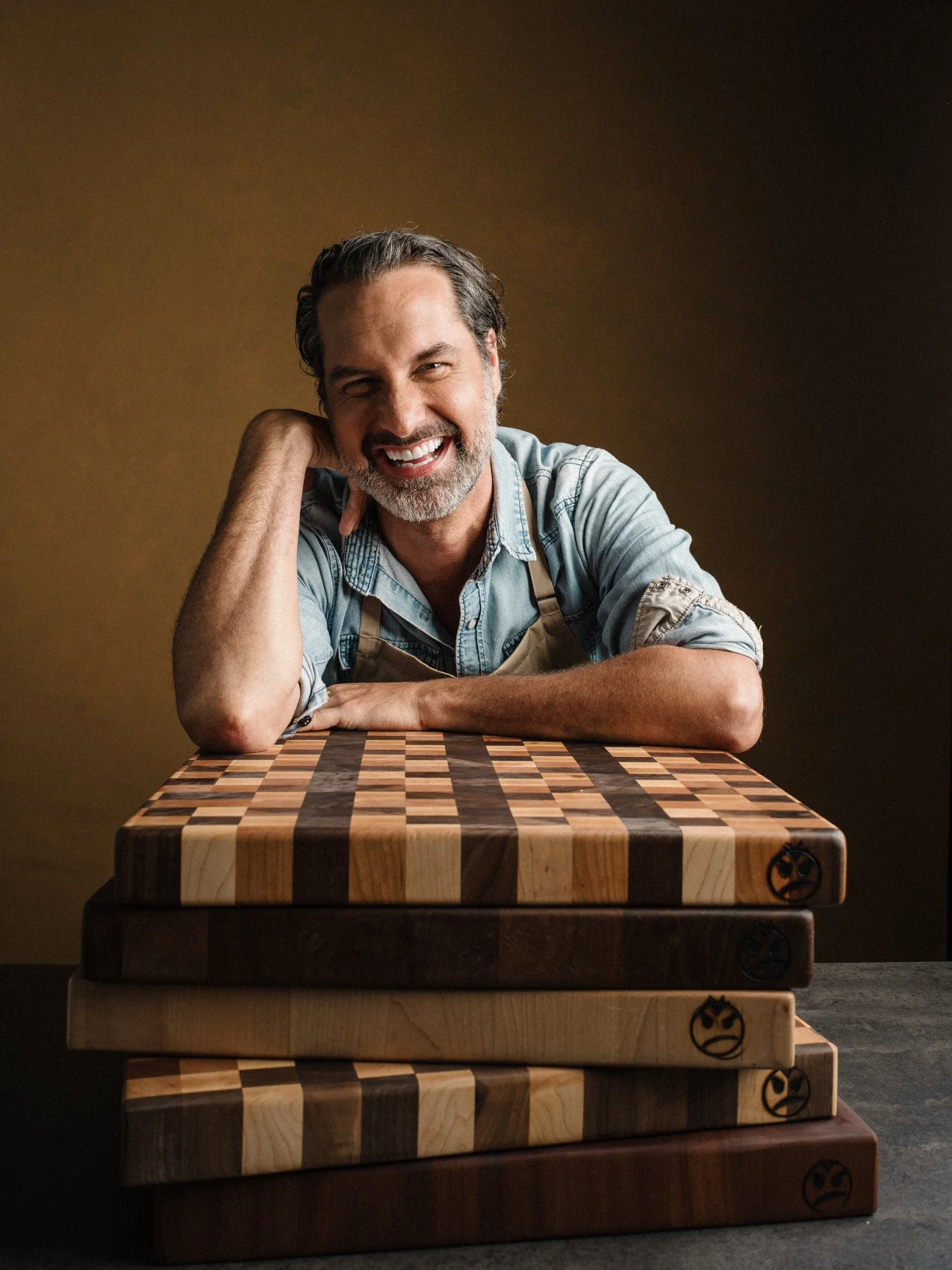 A smiling man with gray hair and beard, wearing a denim shirt and apron, sitting at a wooden table with a chessboard pattern, against a plain brown background.