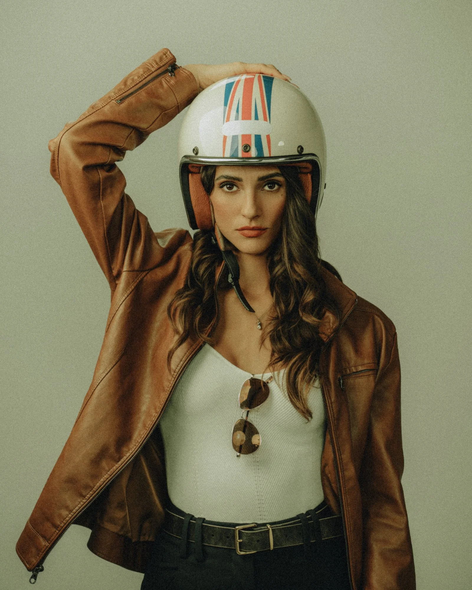 Woman wearing a motorcycle helmet, brown leather jacket, white top, sunglasses hanging from her shirt, and belt, against a plain background.