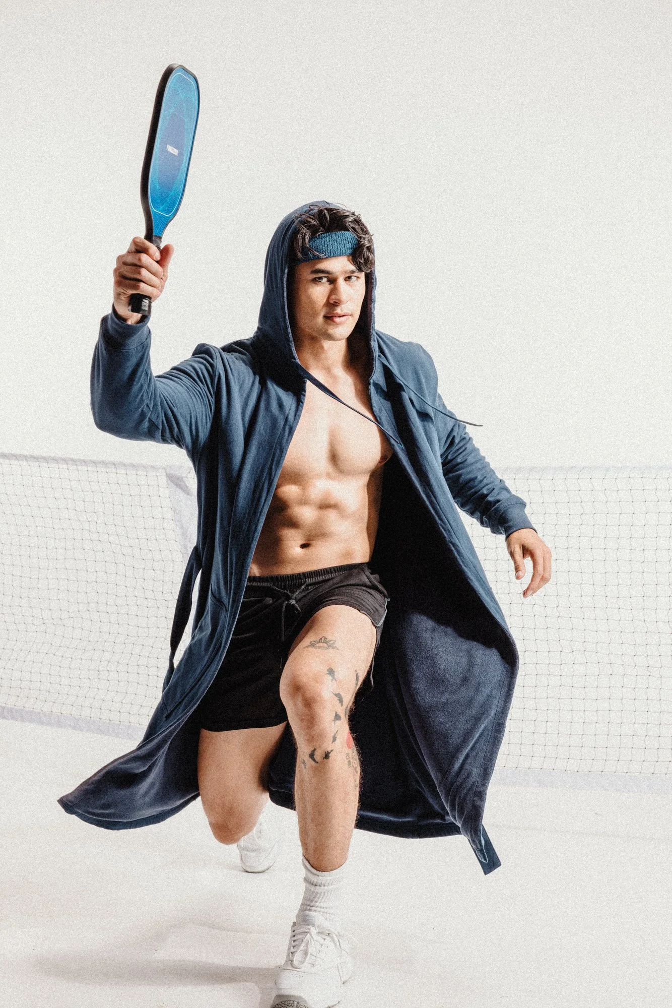 A young man with tattoos on his thigh, wearing white sneakers, white socks, black shorts, a blue hoodie, and a blue headband, holding a tennis racket in a pose on a tennis court with a white net and plain white background.