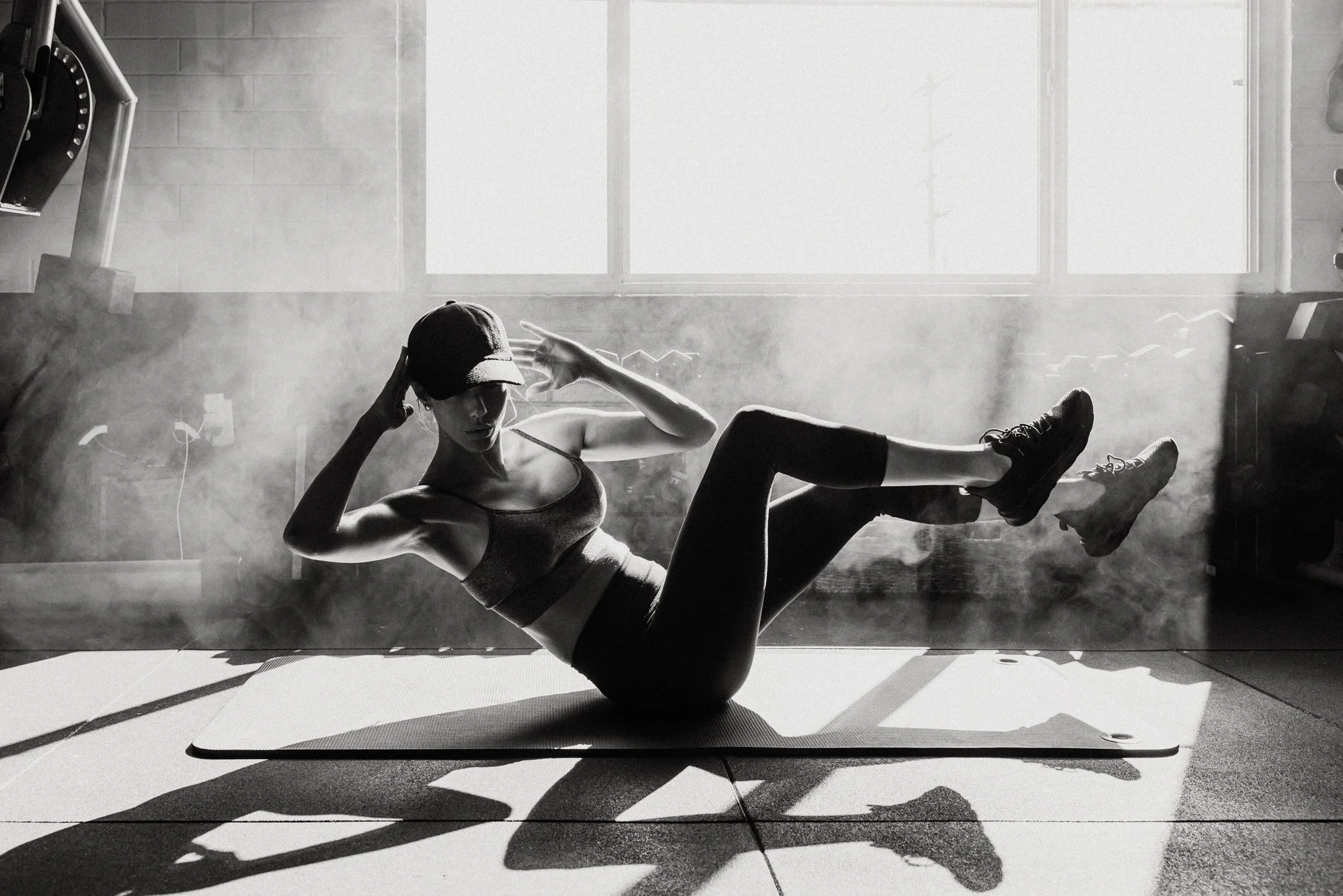 A woman wearing workout clothes and a cap is sitting on a mat in a gym, performing a core exercise with one leg in the air and her hands on her head, with sunlight streaming through windows creating long shadows.
