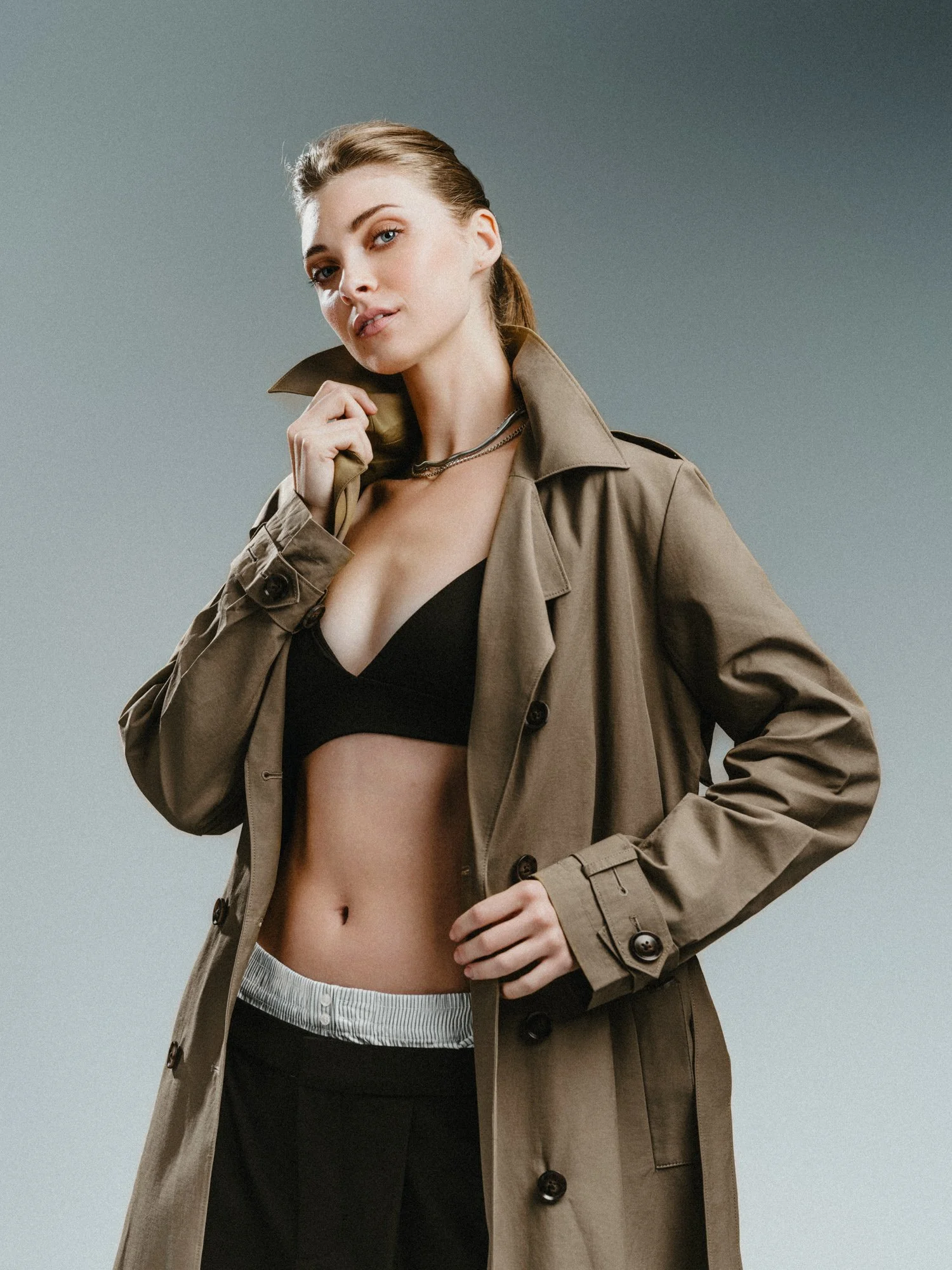 A woman with fair skin, blue eyes, and blonde hair tied back, wearing a beige trench coat over a black crop top, and gray sweatpants, posing with a confident expression against a plain gray background.