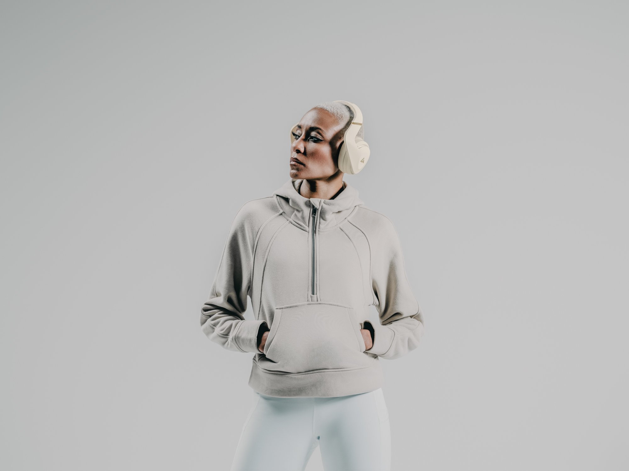 A woman with short, gray hair wearing a beige hoodie with hands in the pockets, standing against a plain gray background, listening to music with over-ear headphones.