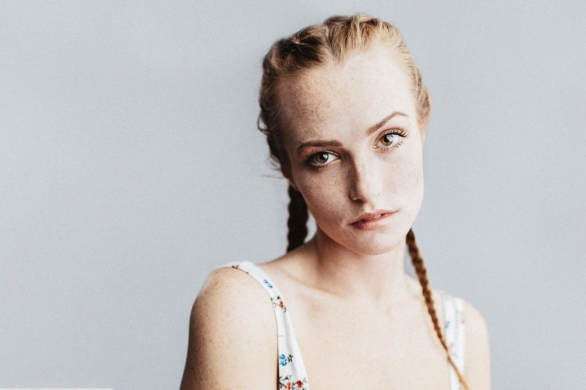 Close-up of a young woman with red hair styled in braids, freckles, and wearing a white floral dress, against a plain gray background.