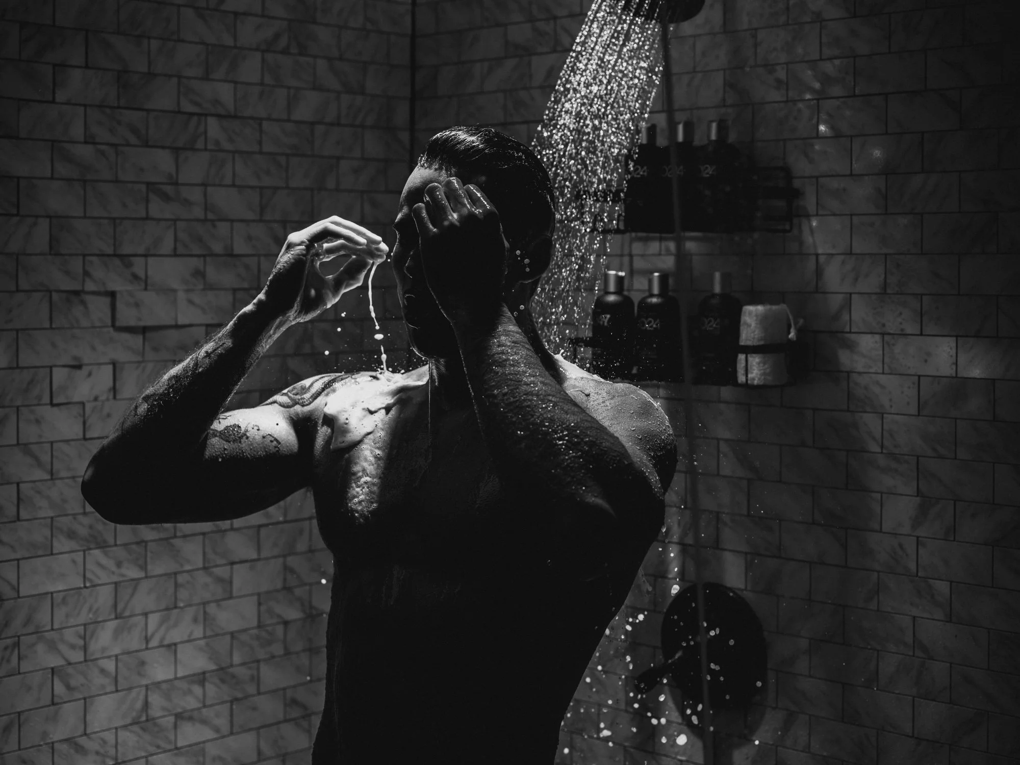 A man is washing himself in a shower with water streaming down from the showerhead. The scene is in black and white, and the bathroom has tiled walls with various bottles and toiletries visible in the background.