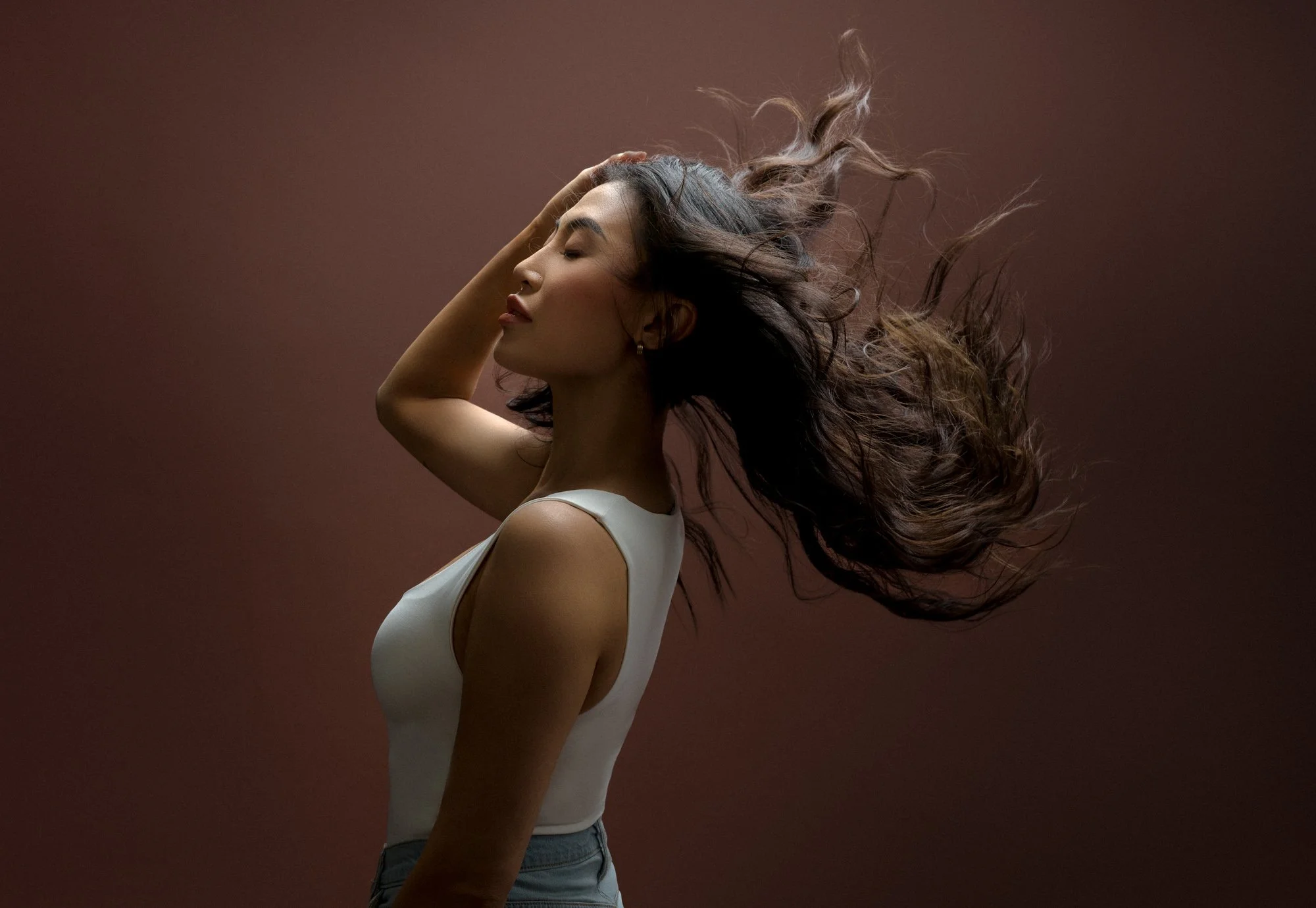 A woman with long curly hair flowing in the air, wearing a sleeveless white top, with her eyes closed and hand resting on her head against a brown background.