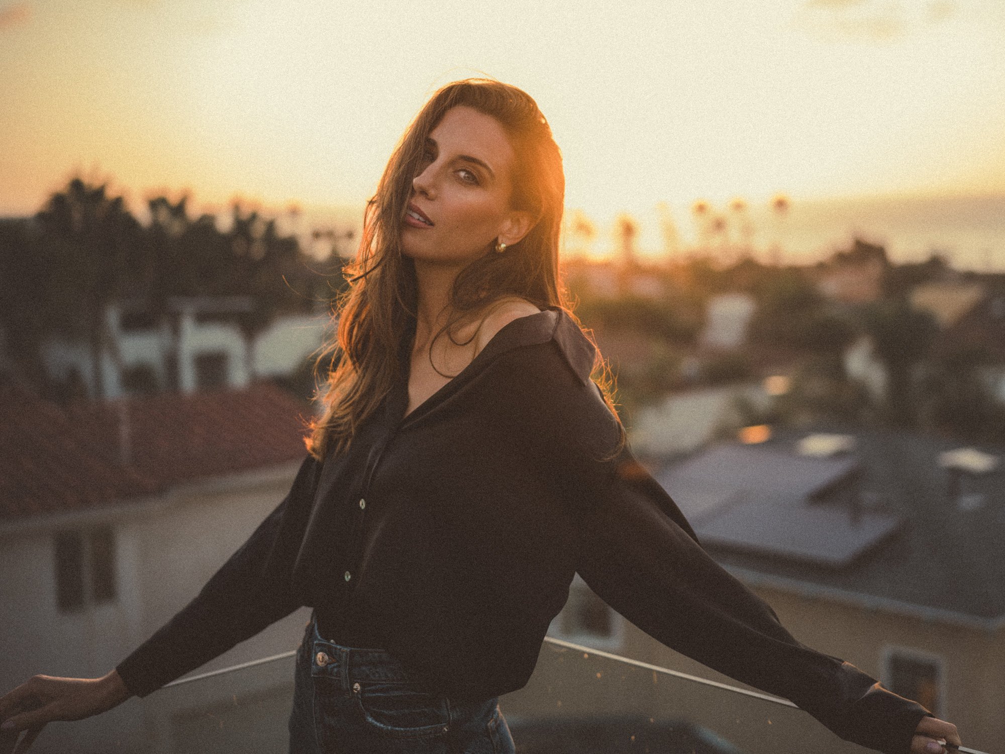 A woman with red hair and blue eyes outdoors during sunset, wearing a black jacket and jeans, leaning on a railing with her arms outstretched.