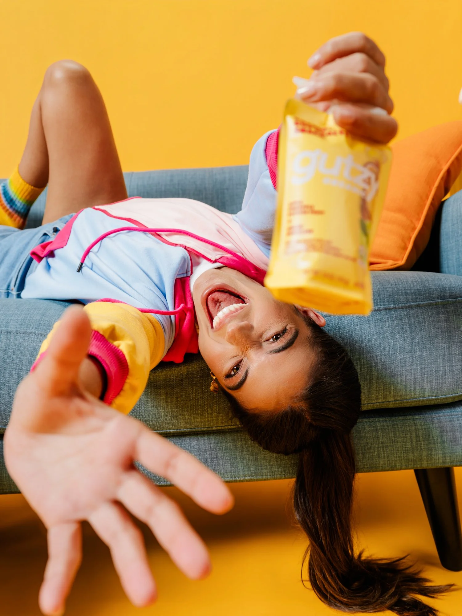 A person lying on a gray couch, smiling, as they hold a bag of potato chips. The background is yellow, and the person is wearing a colorful jacket with rainbow socks.