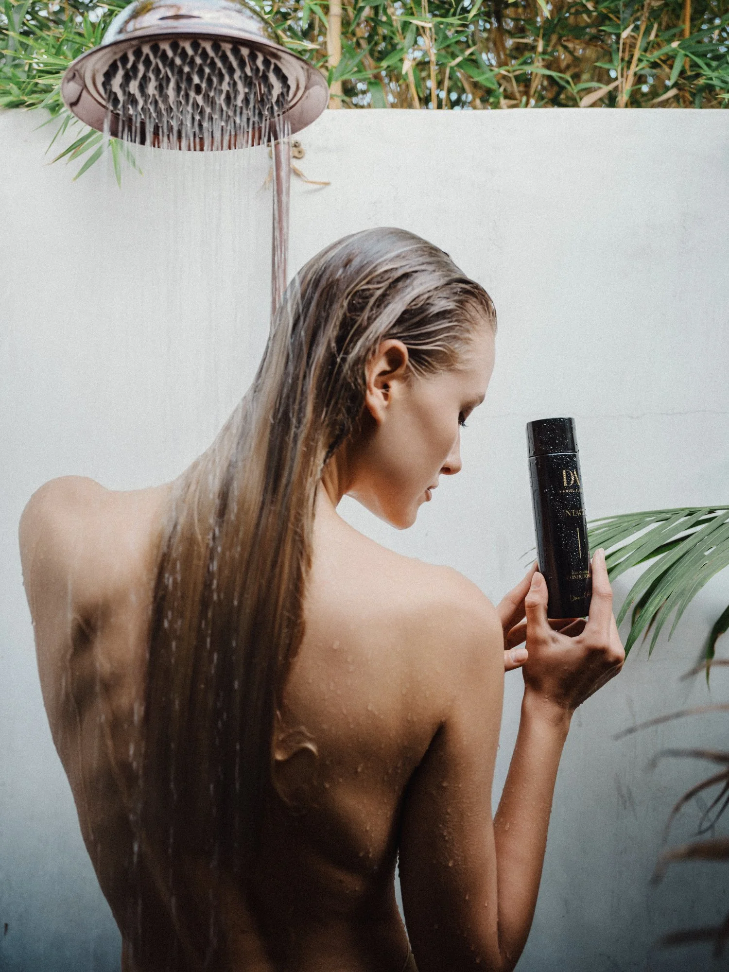 A woman with long wet hair taking a shower outdoors. She is holding a black bottle of hair product, and there are green plants in the background.