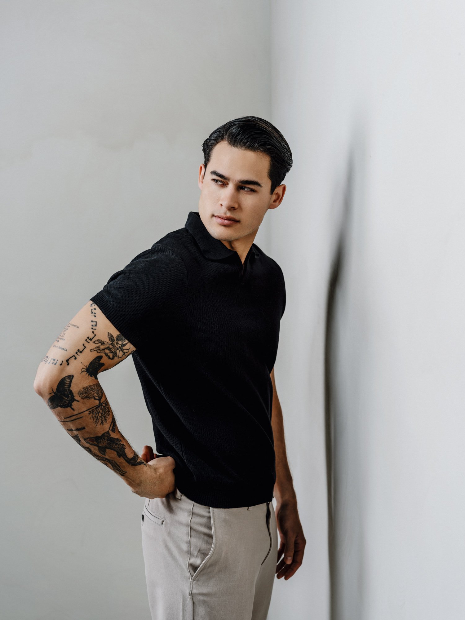 A young man with styled black hair wearing a black polo shirt and light-colored pants, standing against a plain white wall, looking to his left with a contemplative expression.