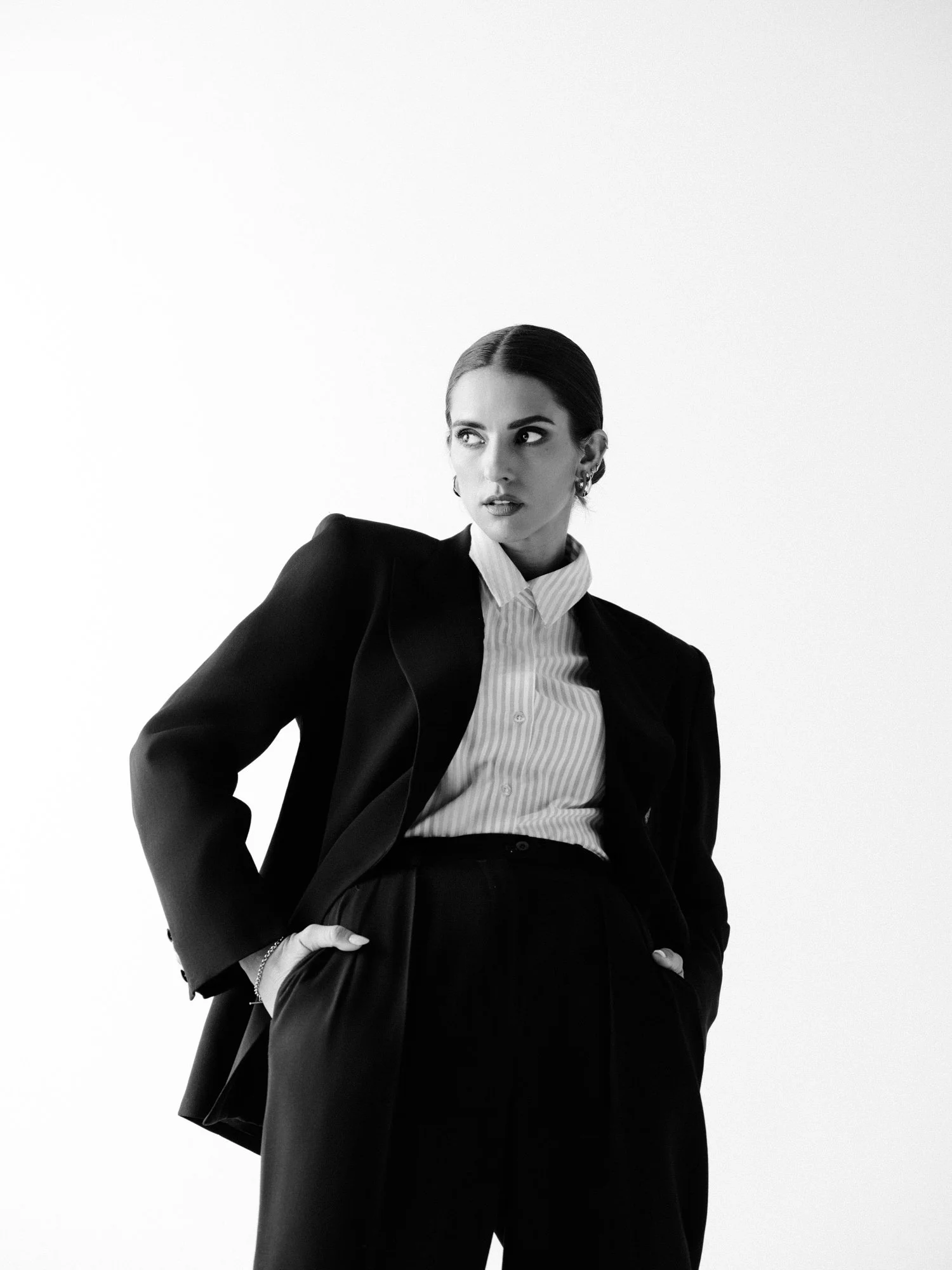 A black-and-white photograph of a woman dressed in a business suit with a collared shirt, standing against a plain white background. She has dark hair slicked back and appears to be looking to her left with a serious expression.