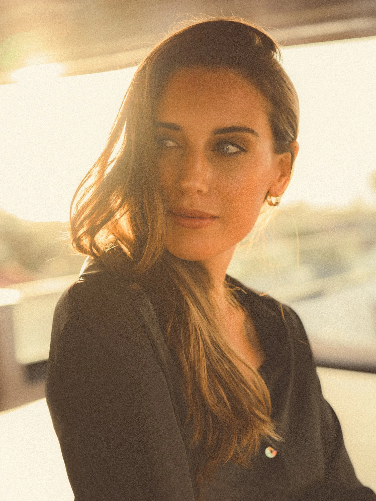 A woman with long, wavy hair and earrings looking to her left, wearing a black top, in front of a softly lit background.