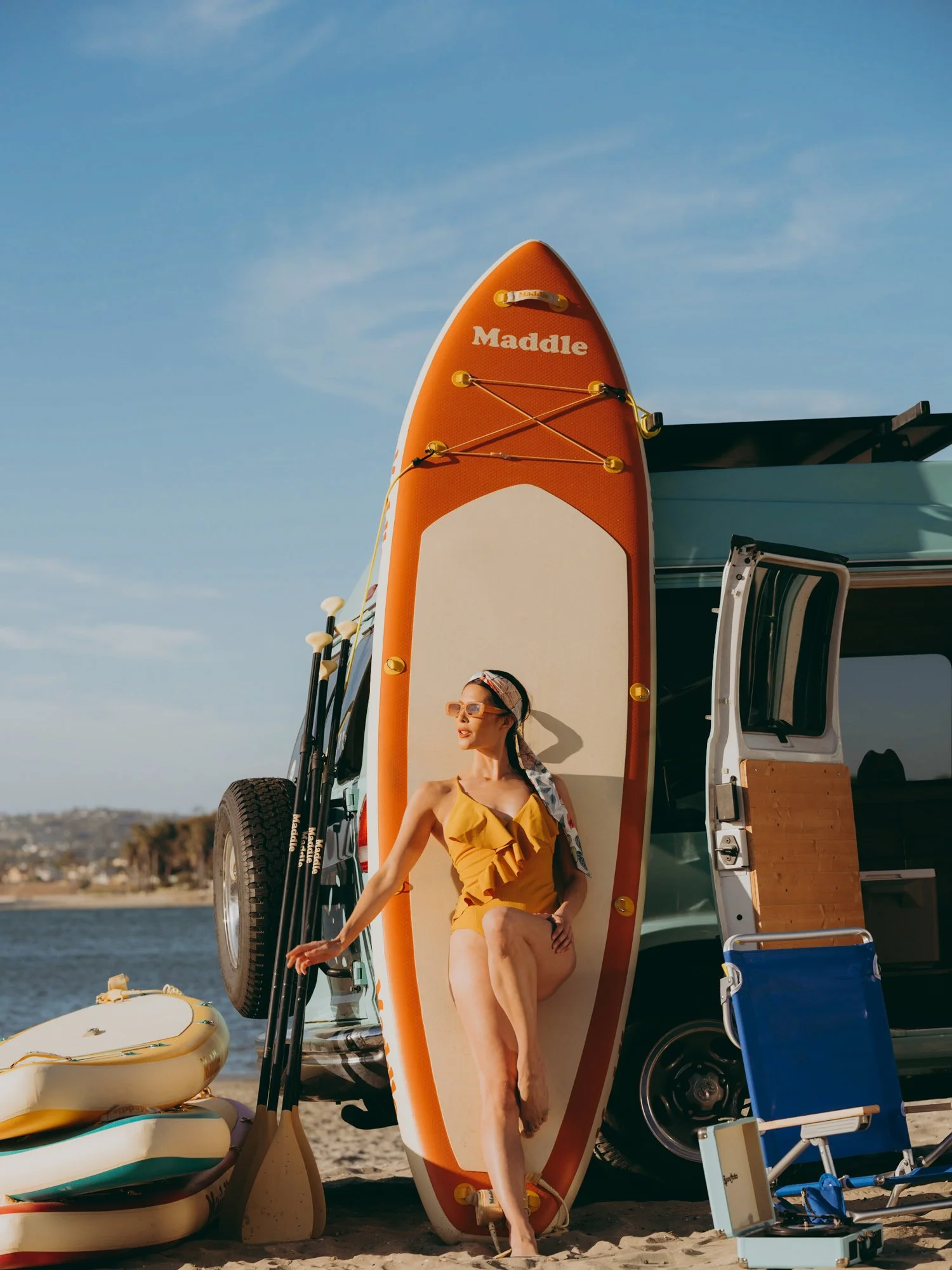 A woman wearing yellow shorts and a matching yellow top sits on a paddleboard next to a vehicle with a standing paddleboard on top. The scene is at a beach near water with a clear sky.