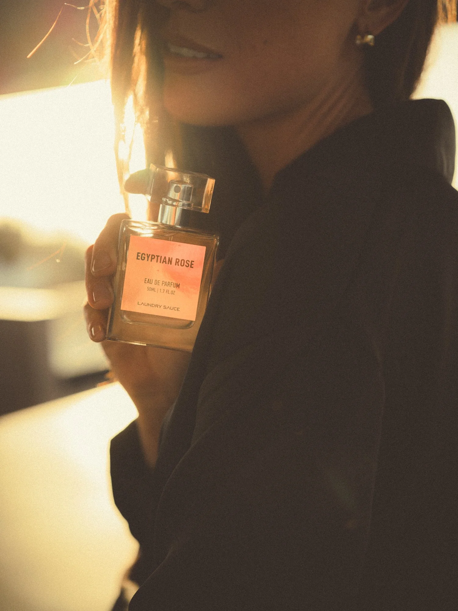 Person holding a bottle of Egyptian Rose perfume, partially visible facing away, wearing a dark jacket, in front of a bright light source.
