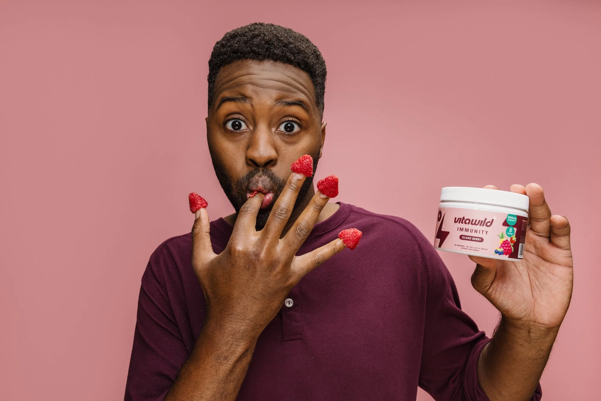 Man holding a container of Vitamin B12 supplements with raspberry flavored gummies falling from his hand, against a pink background.