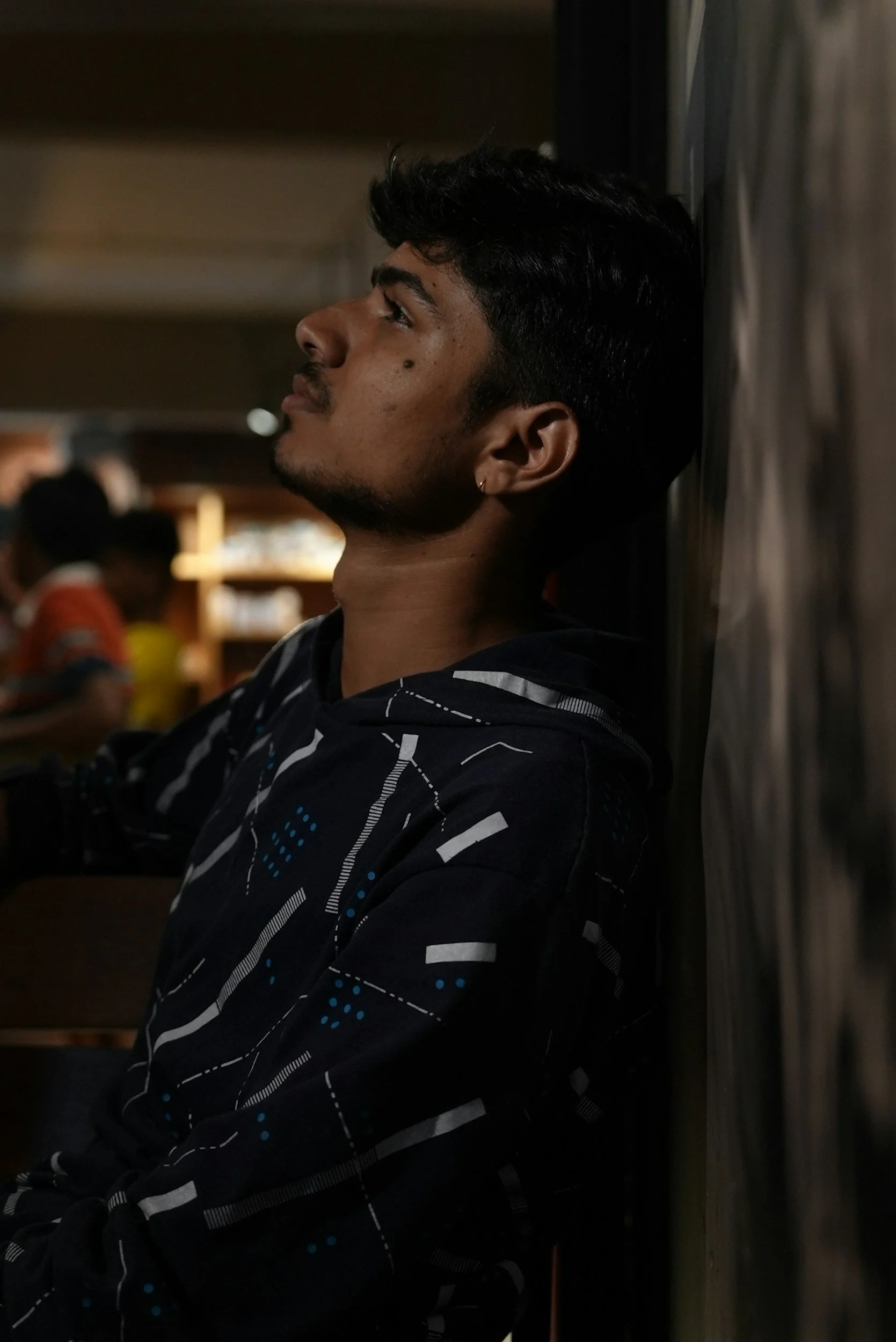 Young man with short, dark hair and medium skin tone sitting against a wall, wearing a black shirt with white and blue patterns, eyes closed or looking upward, in a dimly lit room with blurred figures in the background.