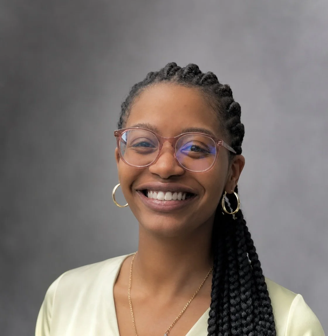A smiling young woman with braids, wearing glasses, hoop earrings, and a light yellow shirt, posed against a gray background.