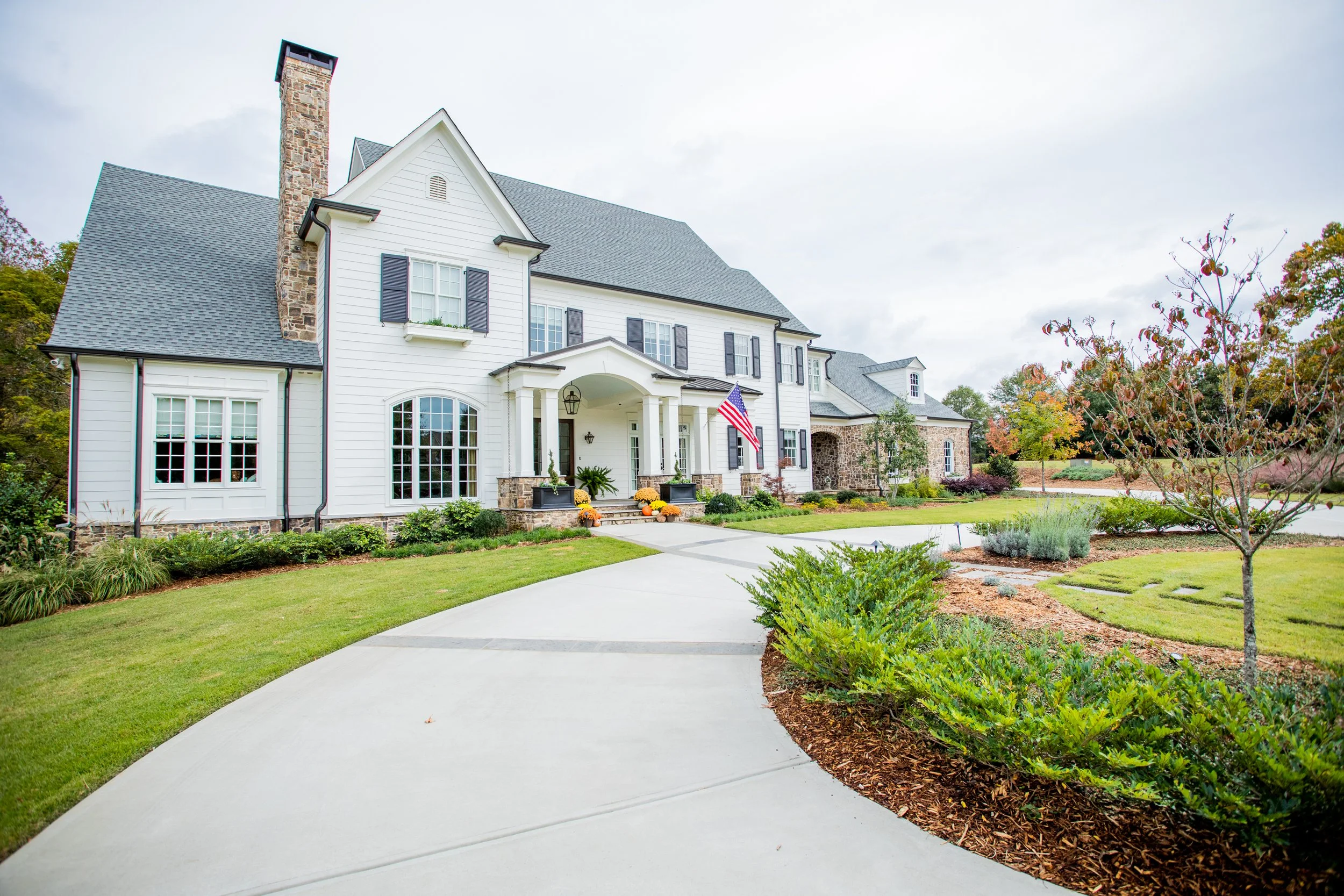 Front view of a large white house with gray roof and stone accents, surrounded by a well-manicured lawn and landscaping, with a curved concrete driveway leading to the front porch.