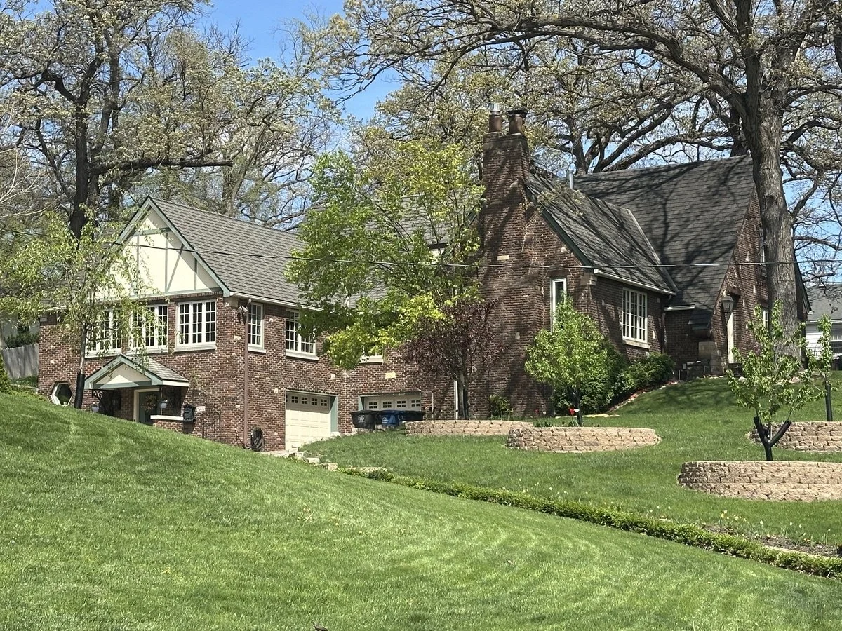 A large brick house with a steep roof, multiple chimneys, and white-framed windows, set on a grassy hill with landscaped plants and trees.