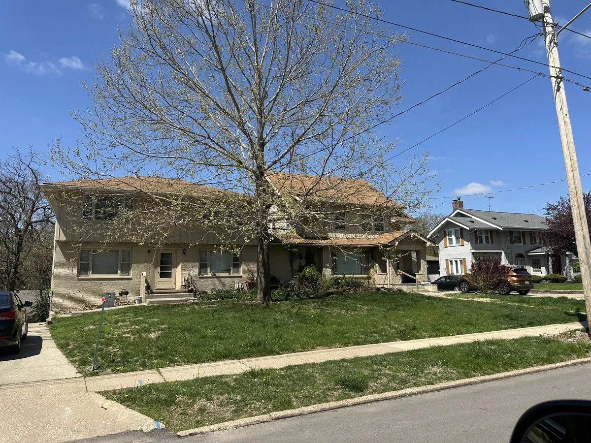 Front view of a two-story residential house with a large tree in the front yard, sidewalk, parked cars, and utility pole on a sunny day with blue sky.