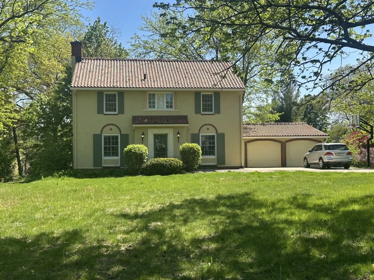 A two-story house with a beige exterior, green shutters, and a red tiled roof, surrounded by trees and a lawn. There is a white SUV parked in the driveway next to a two-car garage, with a basketball hoop nearby.