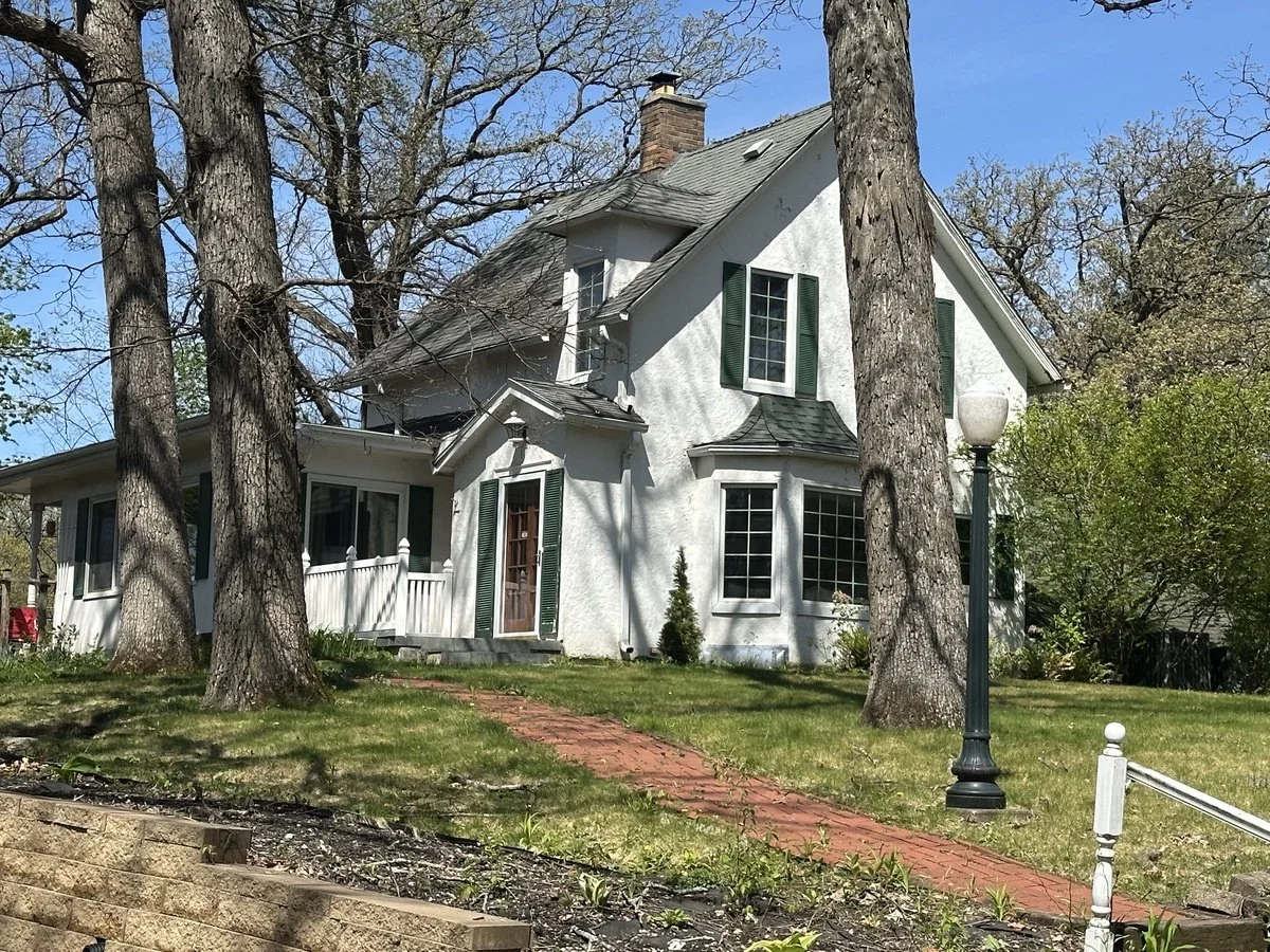A white two-story house with a steep gable roof, surrounded by large trees, with green shutters, a brick pathway leading up to the front door, and a lamp post in the yard.