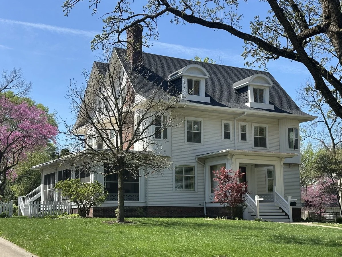 White, three-story house with a dark roof, surrounded by pink flowering trees and a well-maintained lawn.