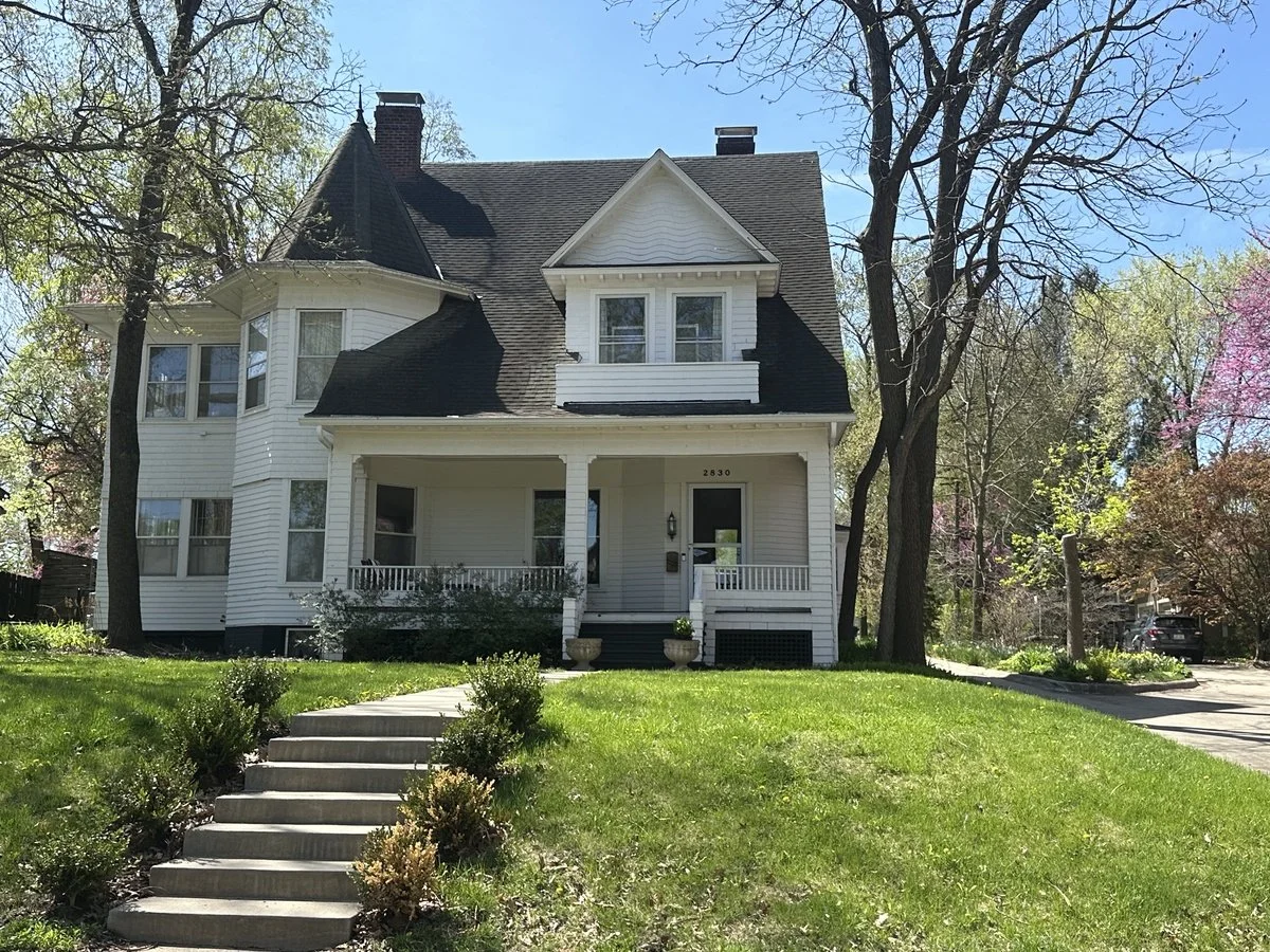 A large white house with a steeply pitched roof and a turret on the left side, surrounded by trees in spring with blooming flowers.