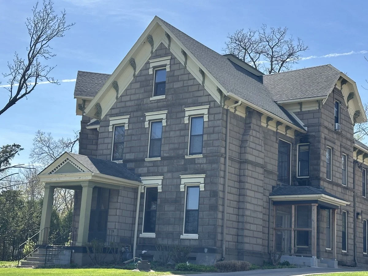 A large three-story Gothic Revival style house made of gray stone with a gabled roof and multiple windows, some with decorative white trim. The house features a small front porch with columns and stairs leading up to it, surrounded by a grassy yard with leafless trees in the background.