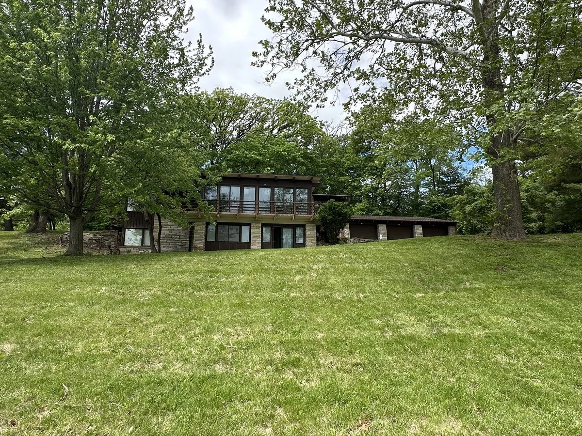 A two-story house with large windows, a balcony, and a garage, surrounded by green trees and grass.