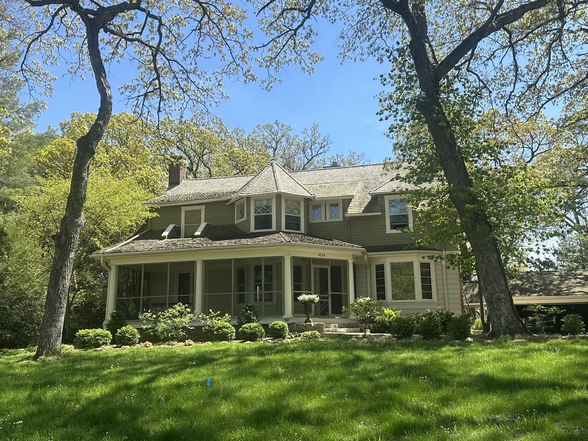 A large, historic house with a porch, surrounded by a well-maintained lawn and trees under a clear blue sky.