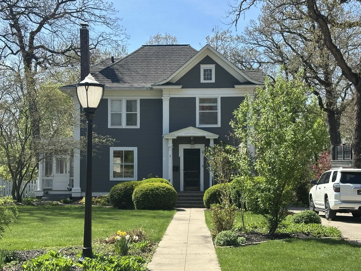 A blue two-story house with white trim, a front porch, and a black front door. The house has a gabled roof and is surrounded by a well-maintained lawn with bushes and trees. A sidewalk leads to the front door, and there is a street lamp near the sidewalk. A white SUV is parked in the driveway on the right.