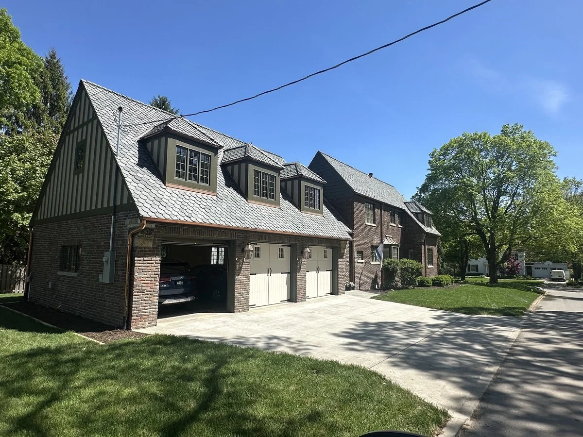 A large suburban house with a brick and wood exterior, multiple dormer windows, a concrete driveway, green lawn, and trees on a clear, sunny day.