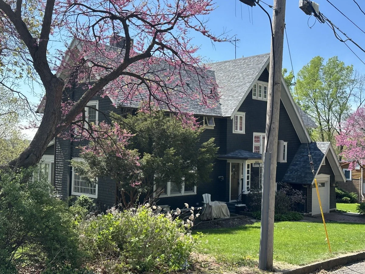 A dark blue house with white trim, surrounded by trees with pink blossoms, green lawns, and a clear blue sky.