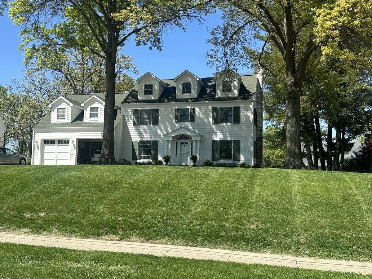 A large, white, multi-story house with black shutters, a prominent front door, and dormer windows on the roof, surrounded by green trees and a well-maintained lawn.