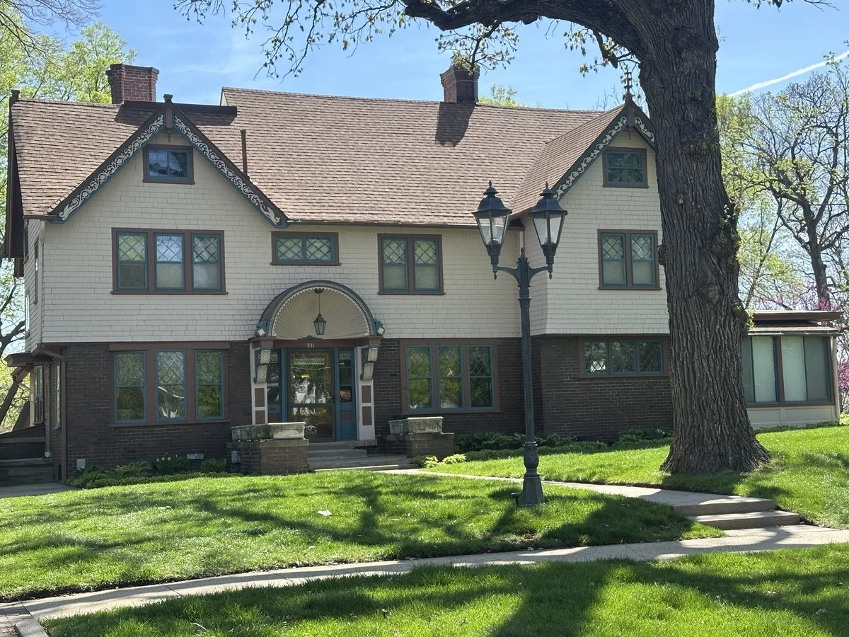 A large, vintage-style house with beige and dark brown brick exterior, multiple windows with diamond-shaped panes, a prominent front door with an arch and lantern above, a street lamp, and a large tree in front with lush green lawn and sidewalk.