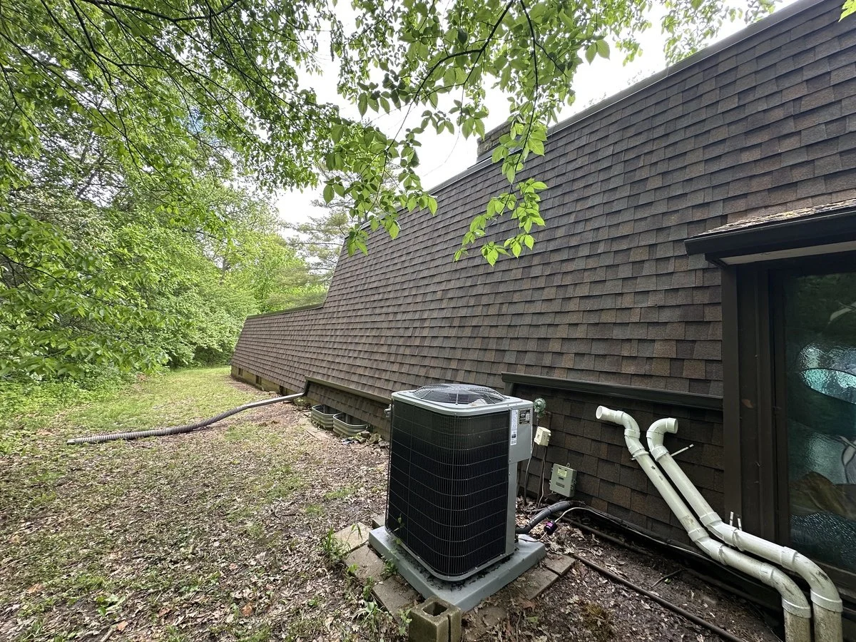 Air conditioning unit installed outside a house with a sloped shingle roof, surrounded by trees.
